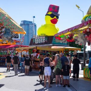 Home Colorful summer carnival scene with a giant rubber duck and game stalls under clear blue sky.